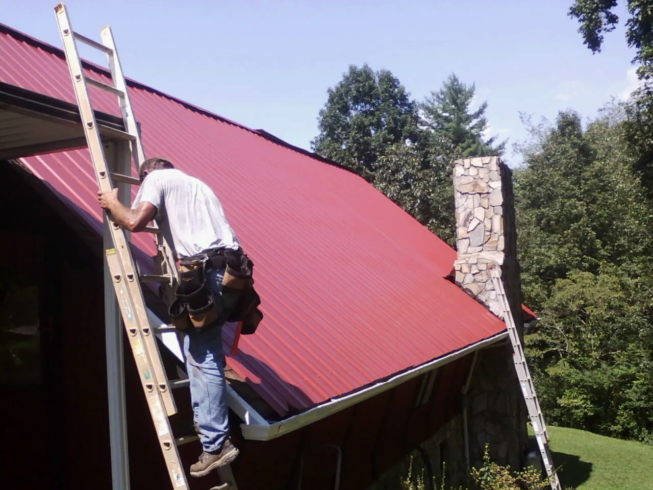 Roofer on a ladder beside a red metal roof and stone chimney — Soffit & Fascia Repair by Roofing Brothers in Grabill